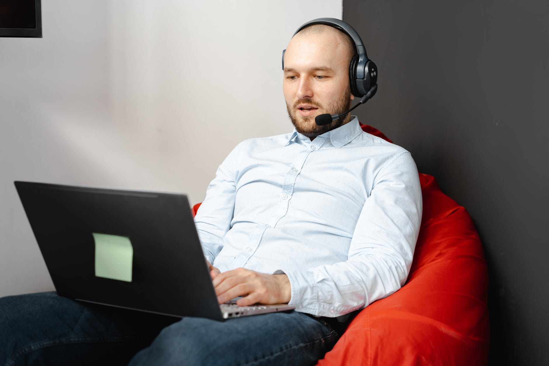 En la imagen aparece un hombre, que porta unos amplios auriculares, sentado en un puff y en cuyas piernas se encuentra un ordenador portátil que está utilizando